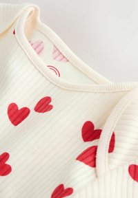 helekreemjas kangas soonilise tekstuuriga, millel on punaste südamekujudega mustrid. Lõpetatud pehme, ümarate kaeluseta ja õmblusdetailidega.