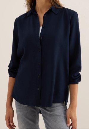 Femme portant une chemise bleu marine boutonnée avec les manches retroussées et un jean gris clair, debout devant un fond neutre.