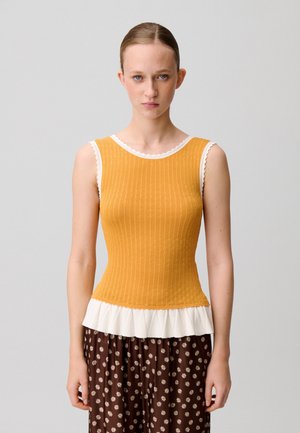 Giovane donna con pelle chiara e capelli raccolti indossa un top senza maniche giallo senape in maglia con orlo a volant bianco e pantaloni marroni a fantasia floreale.