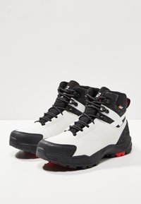 Valkoiset ja mustat vaelluskengät, joissa on nylonia ja nubukkia yläosassa, teksturoitu musta kaulus, mustat nauhat ja punaiset yksityiskohdat pohjassa.