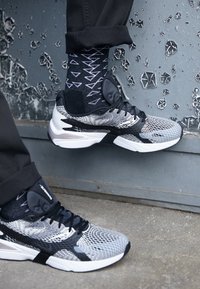 Le scarpe sportive in bianco e nero presentano una tomaia in rete con un motivo geometrico, abbinate a calze nere a fantasia e pantaloni scuri.