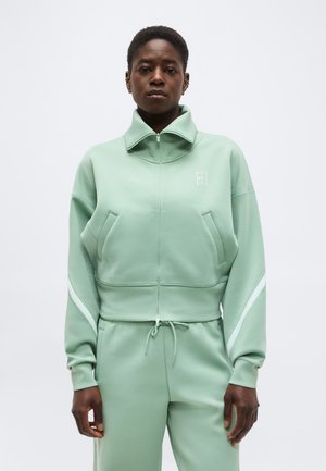 Sort person iført mintgrøn jakke med lynlås og høj krave samt matchende bukser med snøre, stående mod en ensfarvet hvid baggrund.