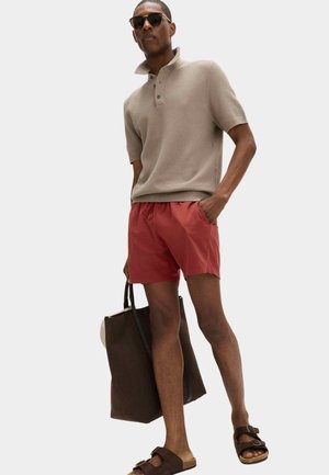 Mand iført beige polo med korte ærmer, røde shorts, brune sandaler og solbriller, holder en stor brun skuldertaske og står med den ene hånd i lommen.