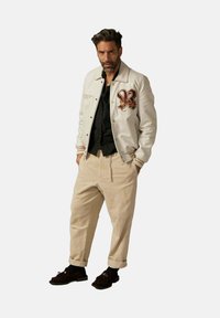 Veste bomber blanche avec des accents brodés, chemise noire, pantalon beige à jambes larges et mocassins marron. Tenue urbaine décontractée avec des textures contrastées.