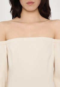 Top cu umeri goi dintr-un material ușor, de culoare crem. Prezintă mâneci elastice și un decolteu simplu, structurat. Textură netedă în întreaga țesătură.