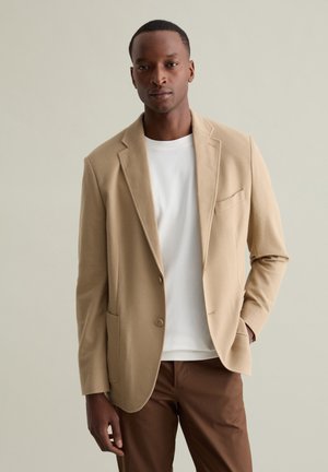 Blazer texturé beige avec une fermeture à bouton unique, des revers crantés et des poches plaquées ; porté sur un t-shirt blanc et un pantalon brun.