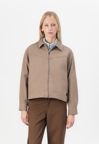 Brown cropped jacket made of thick fabric, featuring a zip front, collar, and two side pockets. Worn over a light blue shirt.