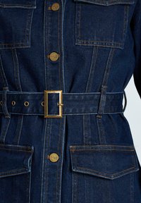 Chaqueta de mezclilla en azul oscuro con herrajes dorados. Presenta grandes bolsillos, una cintura con cinturón y detalles de costura en contraste.