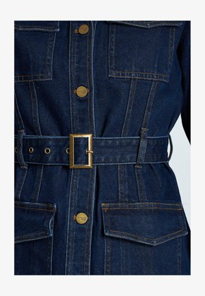 Veste en denim bleu foncé avec quincaillerie dorée. Dispose de grandes poches, d'une taille ceinturée et de détails en surpiqûre contrastée.