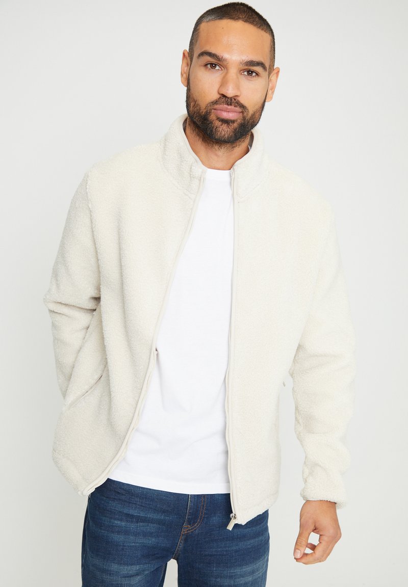 Giacca in pile color crema con colletto alto, chiusura frontale con zip e tasche laterali, caratterizzata da una trama morbida e un fit casual.