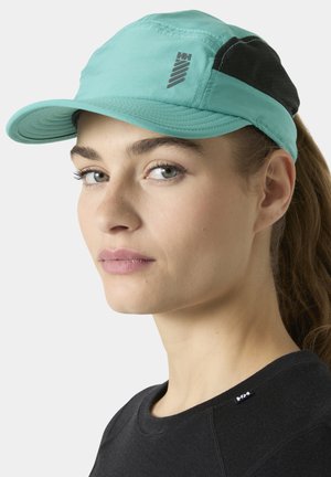 Vrouw met een lichte huid en groene ogen, die een turquoise en zwarte pet draagt en een zwart shirt met een ronde hals en een klein logo vlakbij de kraag.