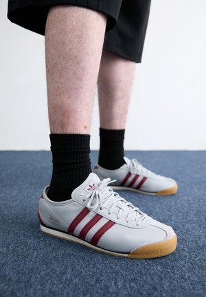 Šedé kožené tenisky s červenými pruhy, gumovou podrážkou a texturovanými detaily. Nosené s černými žebrovanými ponožkami a spárovány s černými šortkami.