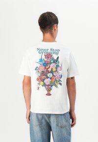 White cotton t-shirt featuring a colorful floral vase graphic on the back, with the text "Never Stop Growing" in blue above the design.