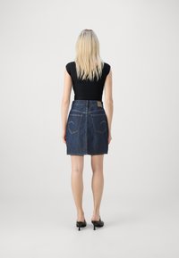 Falda de mezclilla oscura con cinturilla ajustada, dos bolsillos traseros y costura visible. Combinada con una blusa negra ajustada y mules de tacón.