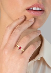 Guldring med en oval rød sten omkranset af små klare sten. Båret på en hånd, med fingre der blidt rører ved ansigtet.