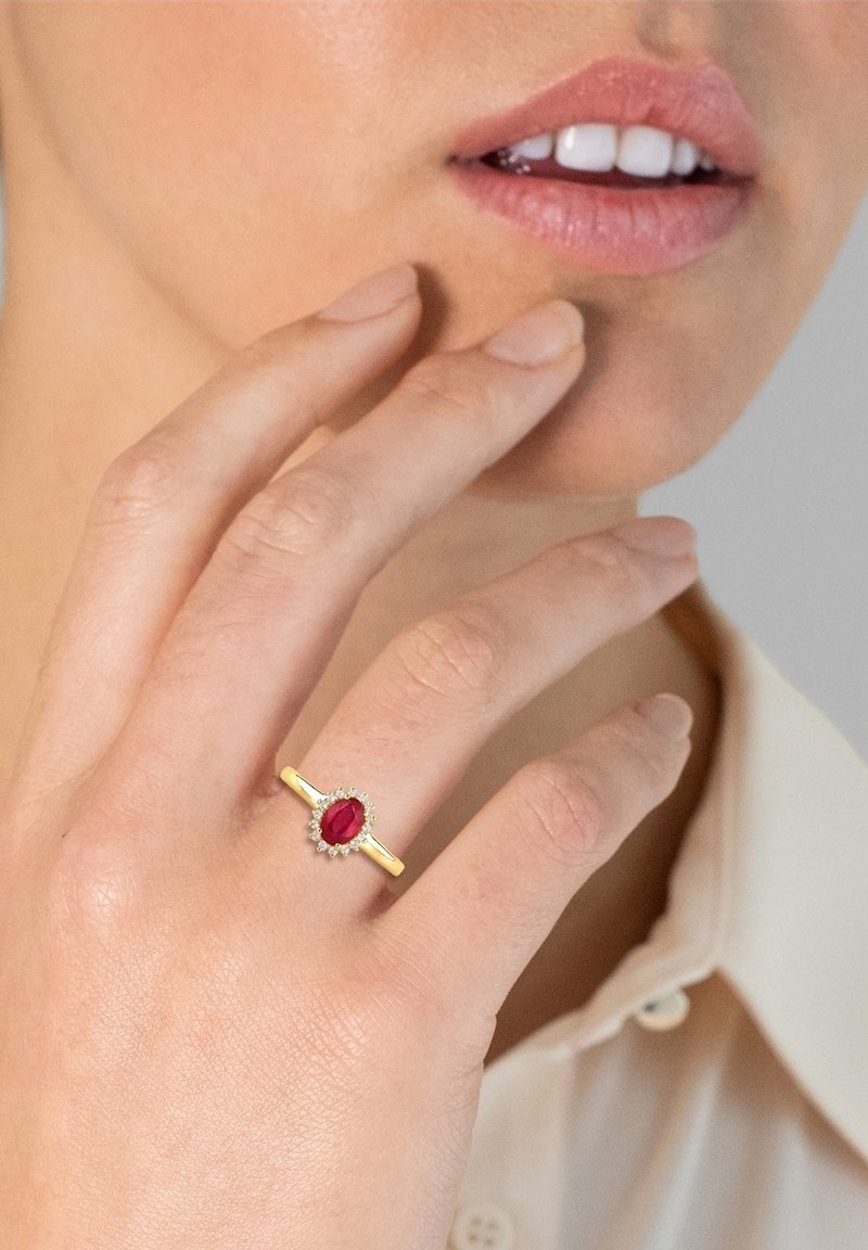 Guldring med en oval rød sten omkranset af små klare sten. Båret på en hånd, med fingre der blidt rører ved ansigtet.