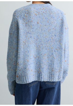 Personne portant un pull texturé bleu clair avec des taches multicolores et un jean bleu foncé, vue de dos.