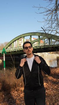 Mladý muž s okuliarmi slnečnými a čiernou bundou si upravuje golier pri zelenom oblúkovom moste nad riekou za jasného počasia.