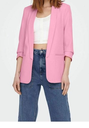 Frau trägt ein pinkes Blazer mit hochgekrempelten Ärmeln über einem weißen Croptop und hochtaillierter, weit geschnittener blauer Jeans, steht mit entspannten Händen.