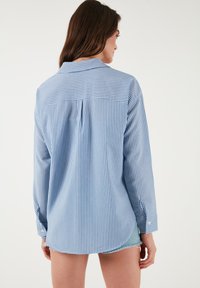 Blue and white striped button-up shirt with long sleeves, curved hem, and a yoke design on the back. Cotton fabric with a slightly relaxed fit.