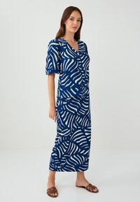 Vestito blu navy con motivo ondulato bianco, scollatura a V, maniche a sbuffo e tasche laterali, realizzato in tessuto leggero che arriva a metà polpaccio, abbinato a sandali marroni.