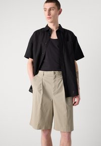 Camisa negra de botones de manga corta sobre una camiseta negra sin mangas, combinada con pantalones cortos beige con pliegues. Diseño simple y tejido ligero y texturizado.