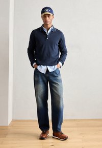 Pullover blu navy con colletto, indossato sopra una camicia azzurro chiaro, abbinato a jeans blu sbiaditi e scarpe marroni con lacci. Cappello con accento azzurro chiaro.