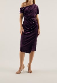 Robe violette en velours, décolletée sur les épaules, avec un design drapé et une coupe à mi-cuisse. Présente une fente latérale et une silhouette ajustée.