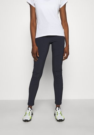 Persoon draagt aansluitende donkerblauwe broek, wit T-shirt met korte mouwen en witte sportschoenen met zwarte en neon groene details.