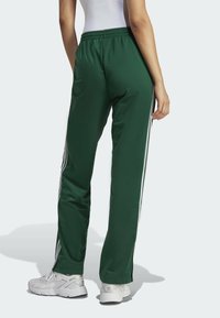 Vihreät urheiluhousut, joissa on sileä pinta, valkoiset sivuraitoja, joustava vyötärö ja rento istuvuus. Päällä on valkoiset tennarit.