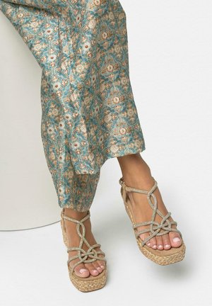 Pieds portant des sandales compensées beiges à lanières avec des semelles tissées, associées à un pantalon ample à motifs floraux bleu-vert.