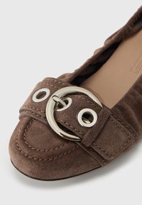 Brun ruskindsflad sko med afrundet tå, bred rem med stor rund metalspænde og dekorative syningsdetaljer.