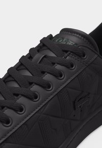 Gros plan d'une basket noire Lacoste avec un motif géométrique texturé, des lacets noirs et un logo crocodile brodé sur le côté.