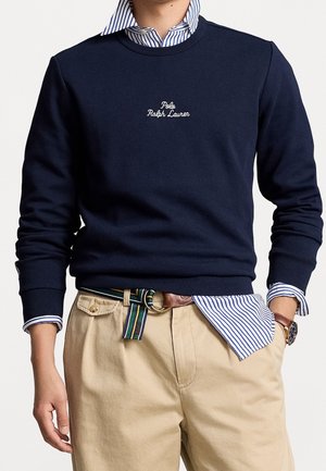 Homme portant un sweat-shirt bleu marine avec le texte « Polo Ralph Lauren » sur une chemise rayée, un pantalon beige, une ceinture rayée et une montre-bracelet avec un bracelet en cuir.