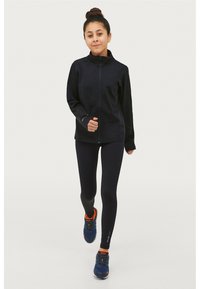 Zwart sportjack met hoge kraag, gecombineerd met zwarte leggings. Draagt blauwe sneakers met oranje accenten en maakt een hardlooppose.