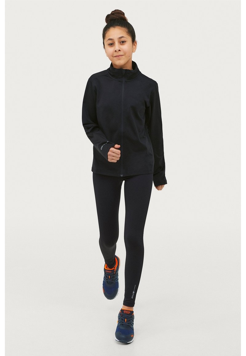 Zwart sportjack met hoge kraag, gecombineerd met zwarte leggings. Draagt blauwe sneakers met oranje accenten en maakt een hardlooppose.