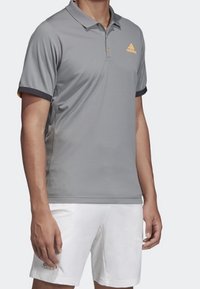 Šedé polo tričko s límečkem, s oranžovým logem adidas na hrudi a kontrastními černými akcenty na rukávech. Nosené s bílými šortkami.