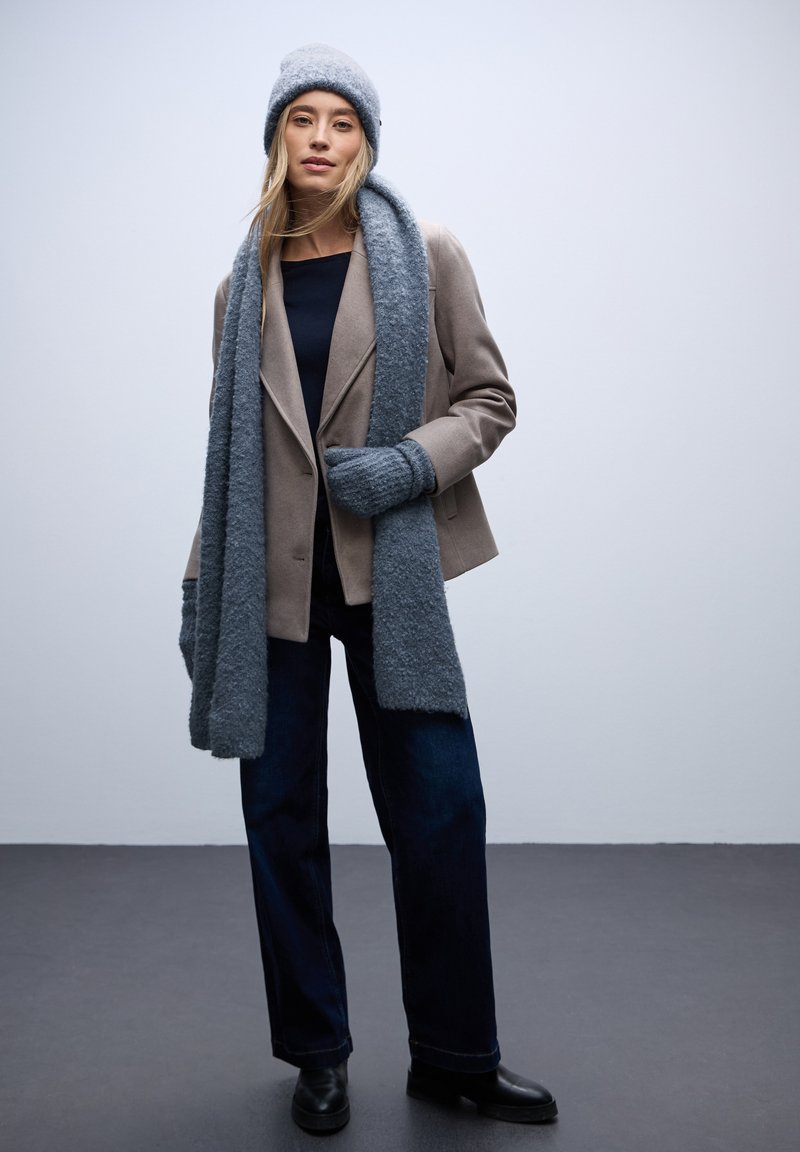 Beżowa marynarka, niebieski dzianinowy szalik, pasująca czapka, szary top, ciemne jeansy wide-leg i czarne botki za kostkę. Miękkie faktury i warstwowy design.