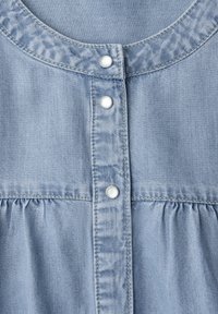 Tissu en denim bleu clair avec trois boutons-pression blancs et des coutures froncées sous la patte de boutonnage sur un vêtement.