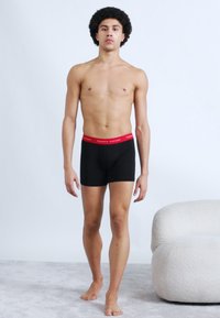 Μαύρα ανδρικά μποξεράκια με κόκκινη μέση, με λεία υφή υφάσματος. Το μοντέλο στέκεται όρθιο μπροστά σε ανοιχτό φόντο.