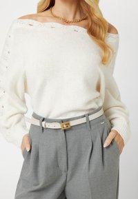 Off-White Strickpullover mit lockerer Passform und weit ausgeschnittenem Halsausschnitt, kombiniert mit grauen, taillierten Hosen und einem hellbeigen Gürtel mit goldener Schnalle.