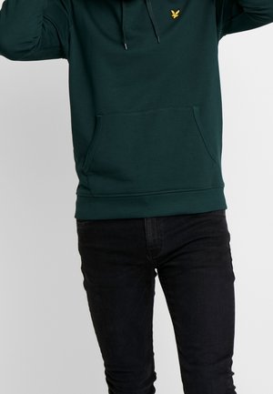 Person iført mørkegrøn hættetrøje med gul fuglelogo og sorte jeans, stående mod en ensfarvet lys grå baggrund.