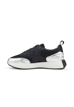 Zapatilla baja negra y plateada con suela blanca, cordones negros y lengüeta en el talón, mostrada de perfil sobre fondo blanco.