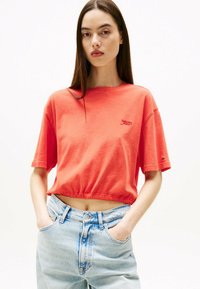 Koralfarvet cropped t-shirt med korte ærmer og rund halsudskæring, der har et lille broderet logo. Bæret med lyseblå højtaljede jeans.