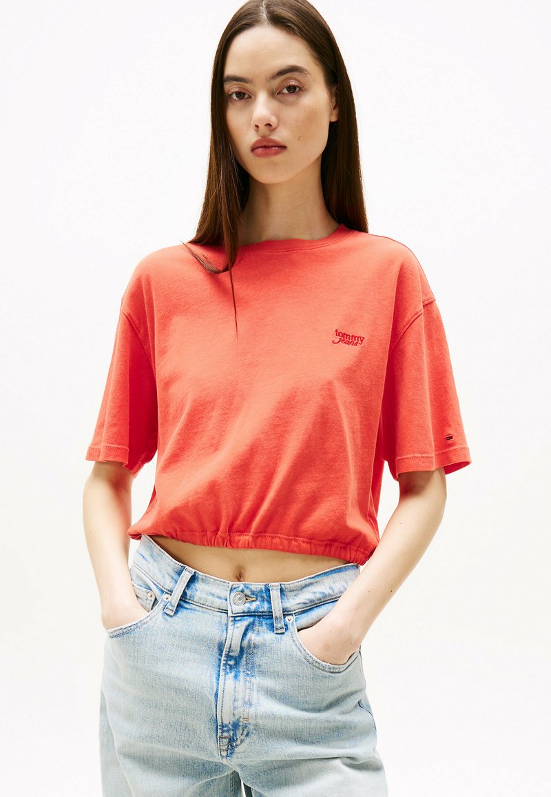 Koralfarvet cropped t-shirt med korte ærmer og rund halsudskæring, der har et lille broderet logo. Bæret med lyseblå højtaljede jeans.