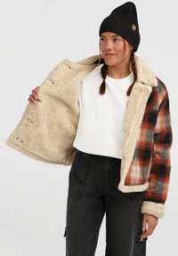 Vrouw die een zwarte beanie, een wit shirt, een zwarte broek en een open bruin en zwart geruit jack met beige schapenvacht voering draagt.