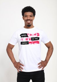 Camiseta de algodón blanca con un diseño gráfico que incluye el texto "ESPÍAS EN LA FAMILIA" y imágenes de ojos en tonos rojos, con un acento negro.