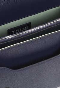 Borsa in pelle viola scuro con esterno texturizzato, chiusura a zip argentata che rivela un interno verde chiaro e un'etichetta nera "TOUS" all'interno.
