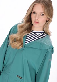 Pelerină impermeabilă teal cu închidere cu capse, cu glugă și buzunar frontal. Purtată peste un tricou cu dungi albastre și albe.