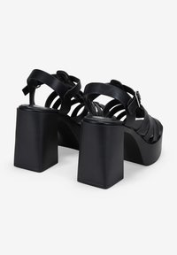 Black platform sandals with wide block heels, featuring crisscross straps and a rear buckle closure. Smooth leather texture and minimalistic design.